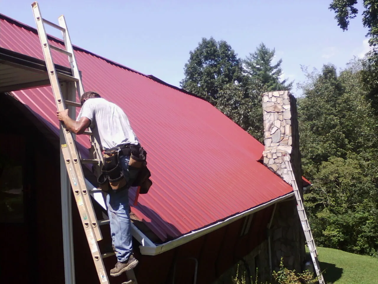 Roofer on a ladder beside a red metal roof and stone chimney — Soffit & Fascia Repair by Roofing Brothers in Cortlandt Manor